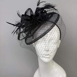 May include: A black fascinator hat featuring a circular base, a structured bow, and delicate feathers. The headpiece is adorned with a fine black netting veil that drapes over the front, all mounted on a thin headband for secure placement.