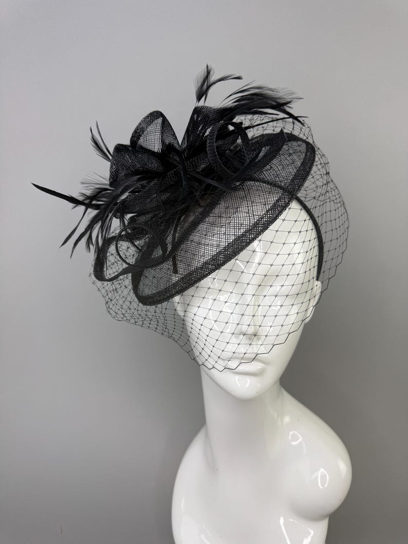 May include: A black fascinator hat featuring a circular base, a structured bow, and delicate feathers. The headpiece is adorned with a fine black netting veil that drapes over the front, all mounted on a thin headband for secure placement.