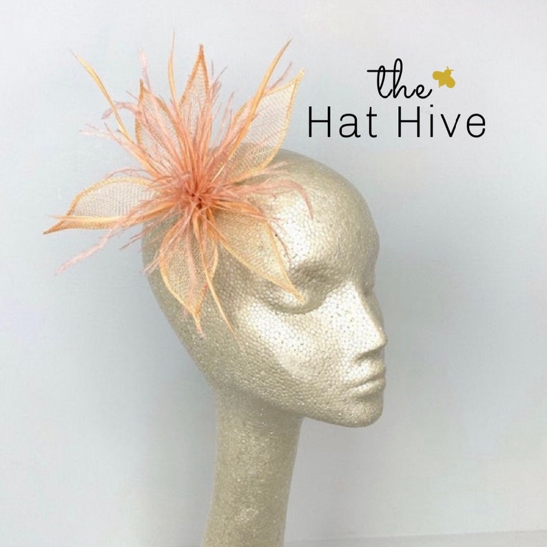 May include: A peach-toned floral headpiece with fine mesh petals and feathery details, presented on a mannequin head. This headpiece is a striking accessory. The text "the Hat Hive" is visible.