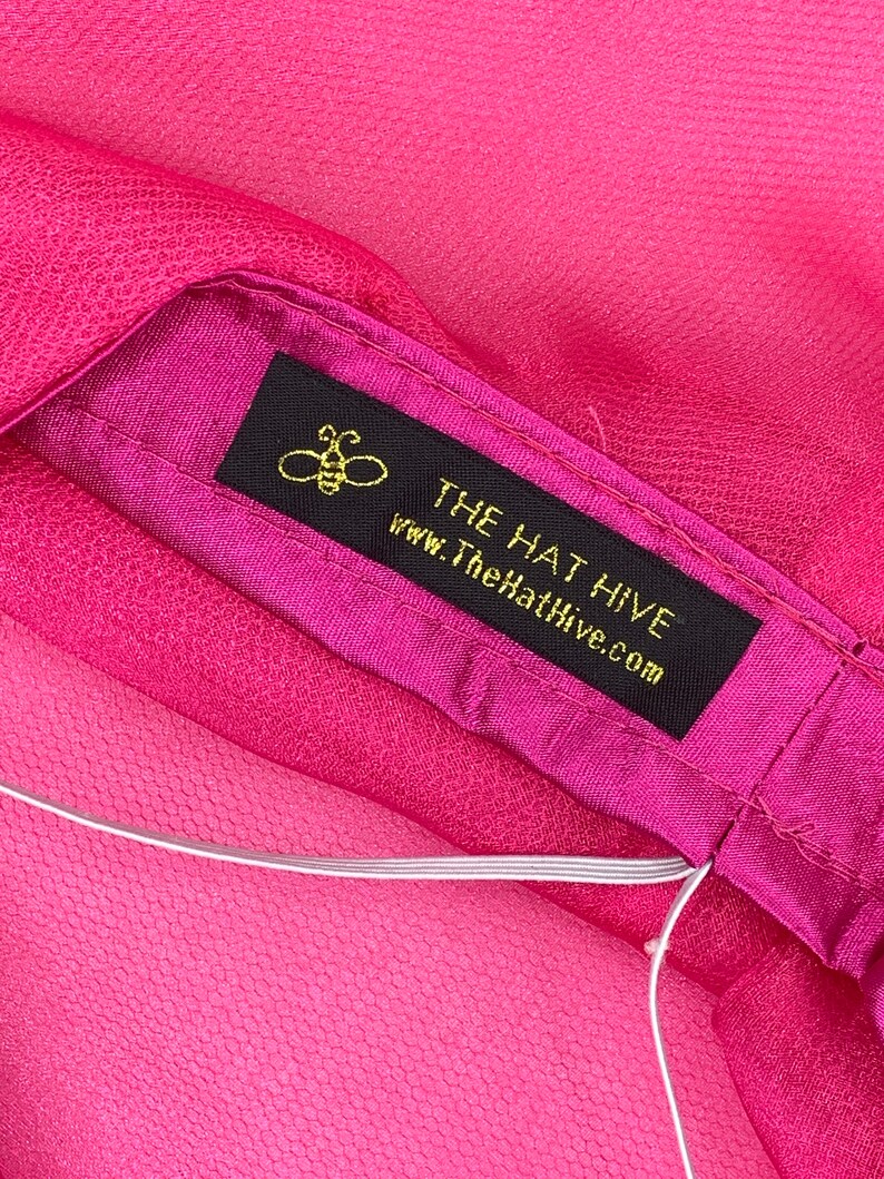 May include: A pink fabric hat with a black label that says "THE HAT HIVE" and "www.TheHatHive.com". A small bee icon is above the text.