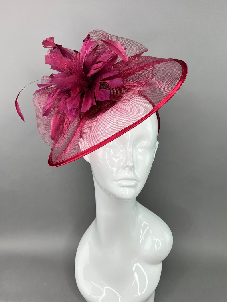 May include: A burgundy fascinator with a large feather flower and a net veil. The fascinator is on a white mannequin head.