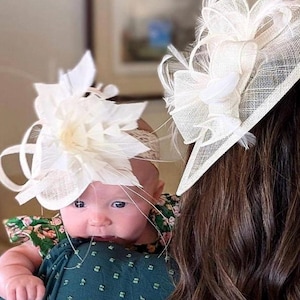 May include: A woman and a baby girl are wearing white hats with feathers. The baby is wearing a green and white floral dress. The woman has long brown hair.