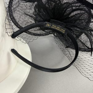 May include: A black fascinator headband featuring a structured mesh bow and a delicate netting veil. The interior of the satin-covered headband includes a black fabric label with gold text that reads THE HAT HIVE and www.TheHatHive.com.