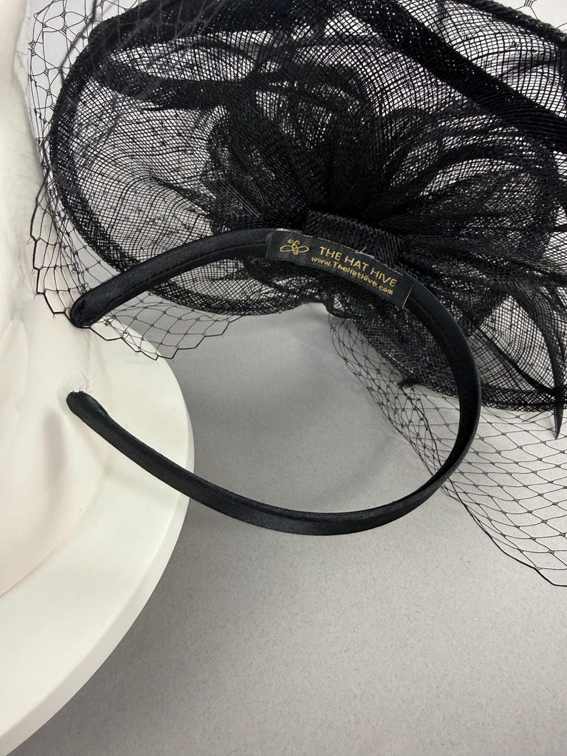 May include: A black fascinator headband featuring a structured mesh bow and a delicate netting veil. The interior of the satin-covered headband includes a black fabric label with gold text that reads THE HAT HIVE and www.TheHatHive.com.