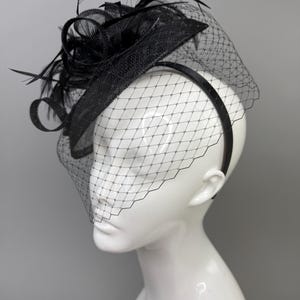 May include: A black fascinator hat featuring a textured, circular base adorned with upright black feathers and loops. A fine black netting veil with a diamond-shaped pattern extends downward from the brim, attached to a thin black headband.
