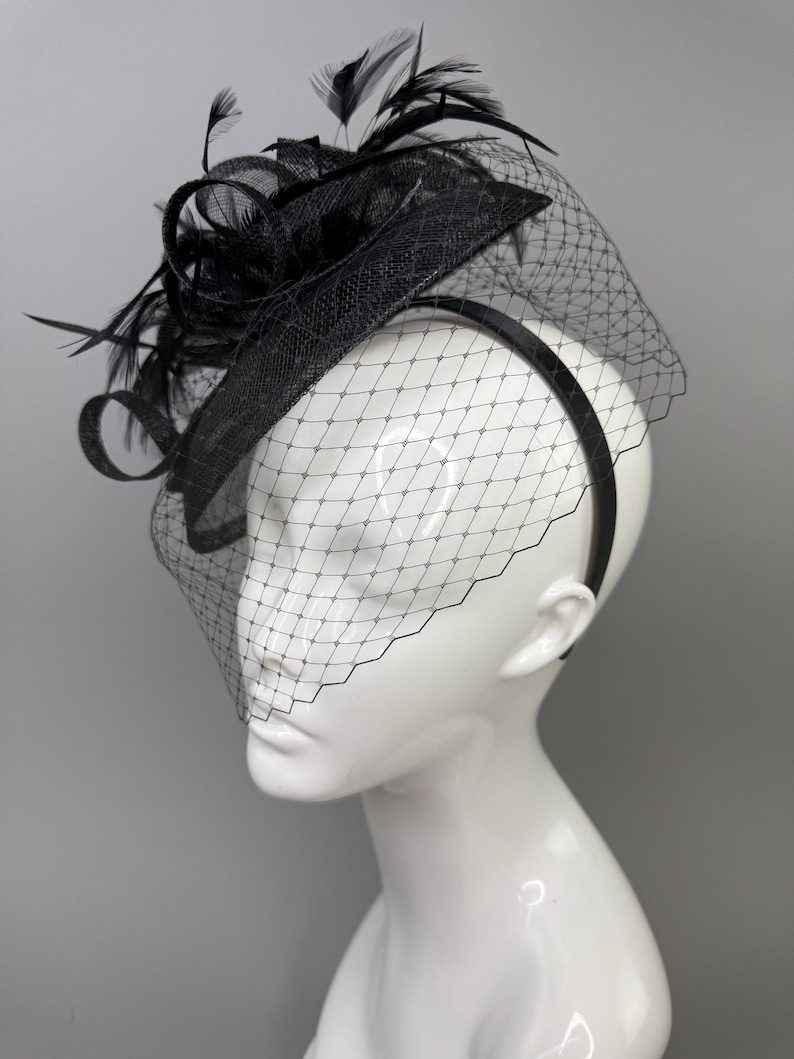 May include: A black fascinator hat featuring a textured, circular base adorned with upright black feathers and loops. A fine black netting veil with a diamond-shaped pattern extends downward from the brim, attached to a thin black headband.