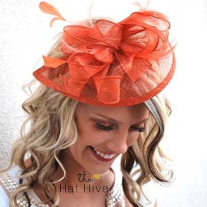 May include: A bright orange fascinator with a large bow and feathers. The fascinator is made of a delicate, netting material.