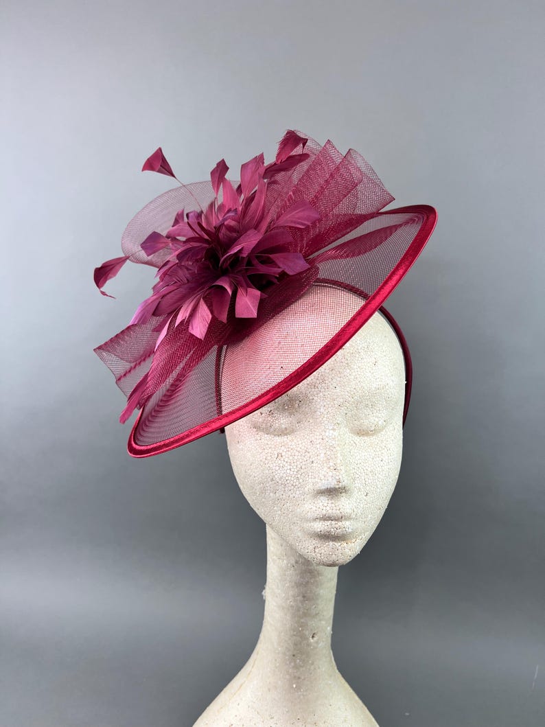 May include: A burgundy fascinator with a large feather and mesh detail. The fascinator is worn on a mannequin's head.
