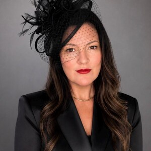 May include: A black fascinator hat featuring a structured base, delicate feathers, and a fine mesh veil that partially covers the face. The headpiece is worn with a black satin blazer and a simple gold chain necklace, creating a formal appearance.