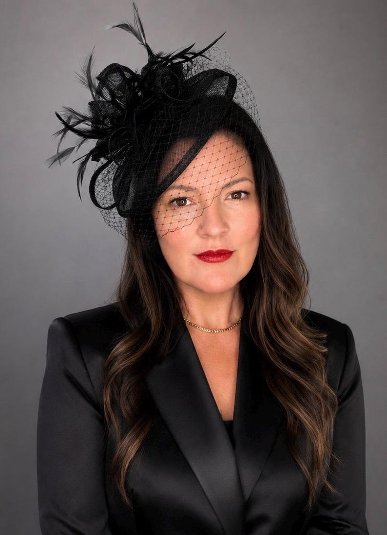 May include: A black fascinator hat featuring a structured base, delicate feathers, and a fine mesh veil that partially covers the face. The headpiece is worn with a black satin blazer and a simple gold chain necklace, creating a formal appearance.