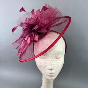 May include: A burgundy fascinator with a large feather and net detail. The fascinator is on a white mannequin head.