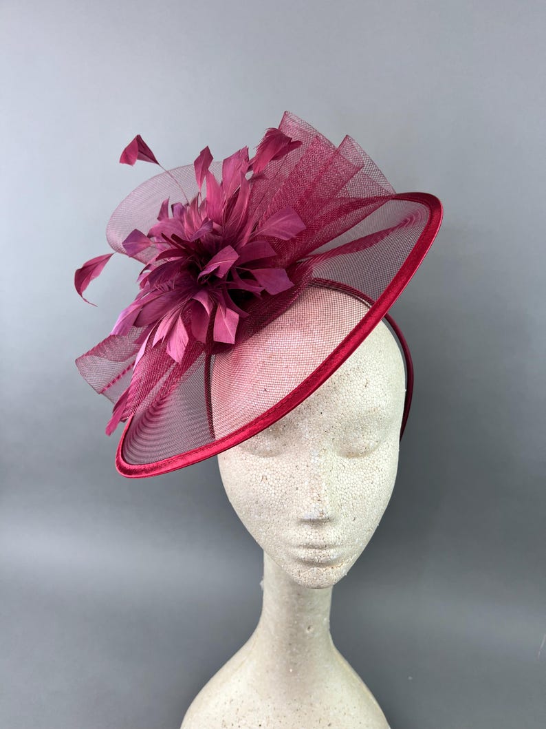 May include: A burgundy fascinator with a large feather and net detail. The fascinator is on a white mannequin head.