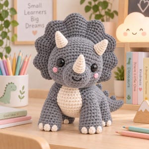 May include: A gray and cream-colored crochet triceratops toy with a large frill, three horns, and black safety eyes. The dinosaur has a textured yarn surface, pink embroidered cheeks, and a small smile, standing on four legs with cream-colored claws.