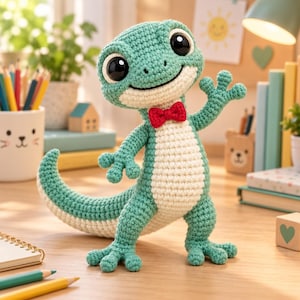 May include: A teal and cream-coloured crocheted lizard plush toy featuring large black glossy eyes, a wide smiling mouth, and a small red crocheted bow tie. The creature stands on two legs with a long curved tail and one arm raised in a waving gesture.