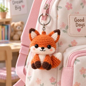 May include: A crochet fox keychain with orange, white, and dark brown yarn, featuring black safety eyes and a small smile, attached to a silver metal ring. It hangs from a light pink backpack with a floral pattern and a patch reading Good Days.