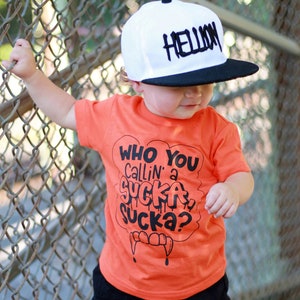 Pu&ograve; includere: Un bambino piccolo che indossa una maglietta arancione con il testo "Who You Callin' a Sucka Sucka?" in nero. Il bambino indossa anche un berretto da baseball nero e bianco con la parola "Hellion" scritta in bianco. Il bambino &egrave; in piedi su una superficie di legno con una recinzione a maglie in background.