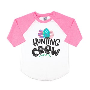以下が含まれることがあります： カラフルなイースターエッグと「Hunting Crew」の文字が描かれた白とピンクのベースボールTシャツです。