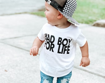 May include: A toddler wearing a white t-shirt with the text "Bad Boy For Life" in black, blue ripped jeans, black sneakers, and a black and white checkered trucker hat.