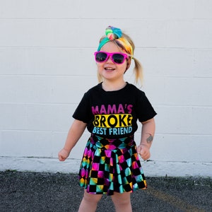 May include: A young girl wearing a black t-shirt with the text "Mama's Broke Best Friend" in colourful letters, a rainbow tie-dye headband, pink sunglasses, and a black and rainbow checkered skirt.