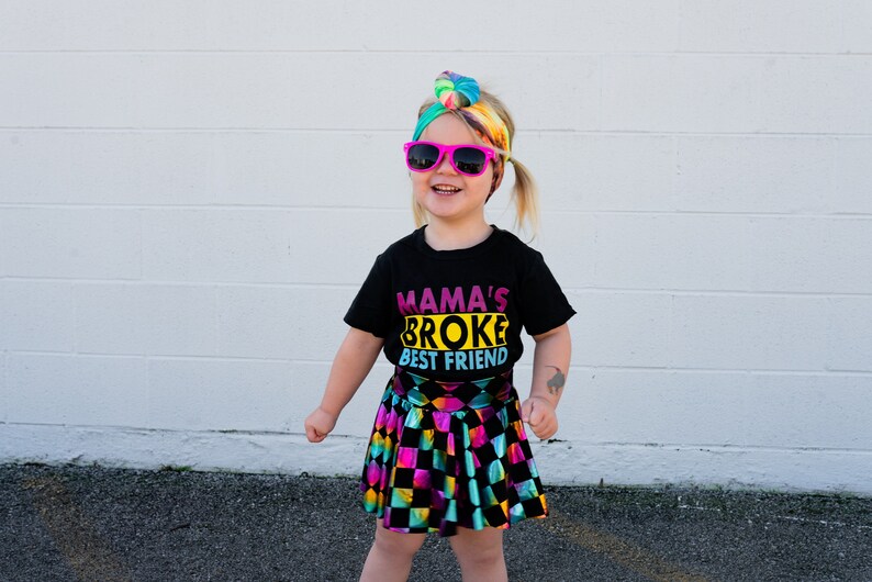 May include: A young girl wearing a black t-shirt with the text "Mama's Broke Best Friend" in colourful letters, a rainbow tie-dye headband, pink sunglasses, and a black and rainbow checkered skirt.
