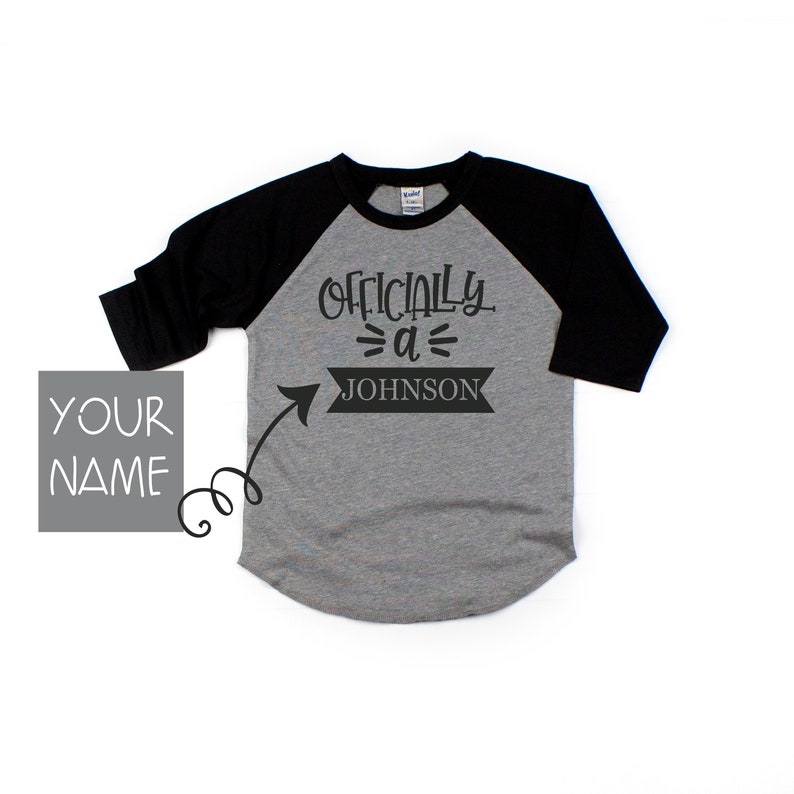 Puede incluir: Una camiseta de b&eacute;isbol gris y negra con el texto "Officially a JOHNSON" en la parte delantera. El texto est&aacute; en una pancarta negra con una flecha que apunta hacia la derecha.
