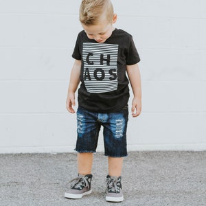 May include: A young boy wearing a black t-shirt with the word "CHAOS" printed in white on it. He is also wearing blue denim shorts and gray high-top sneakers.