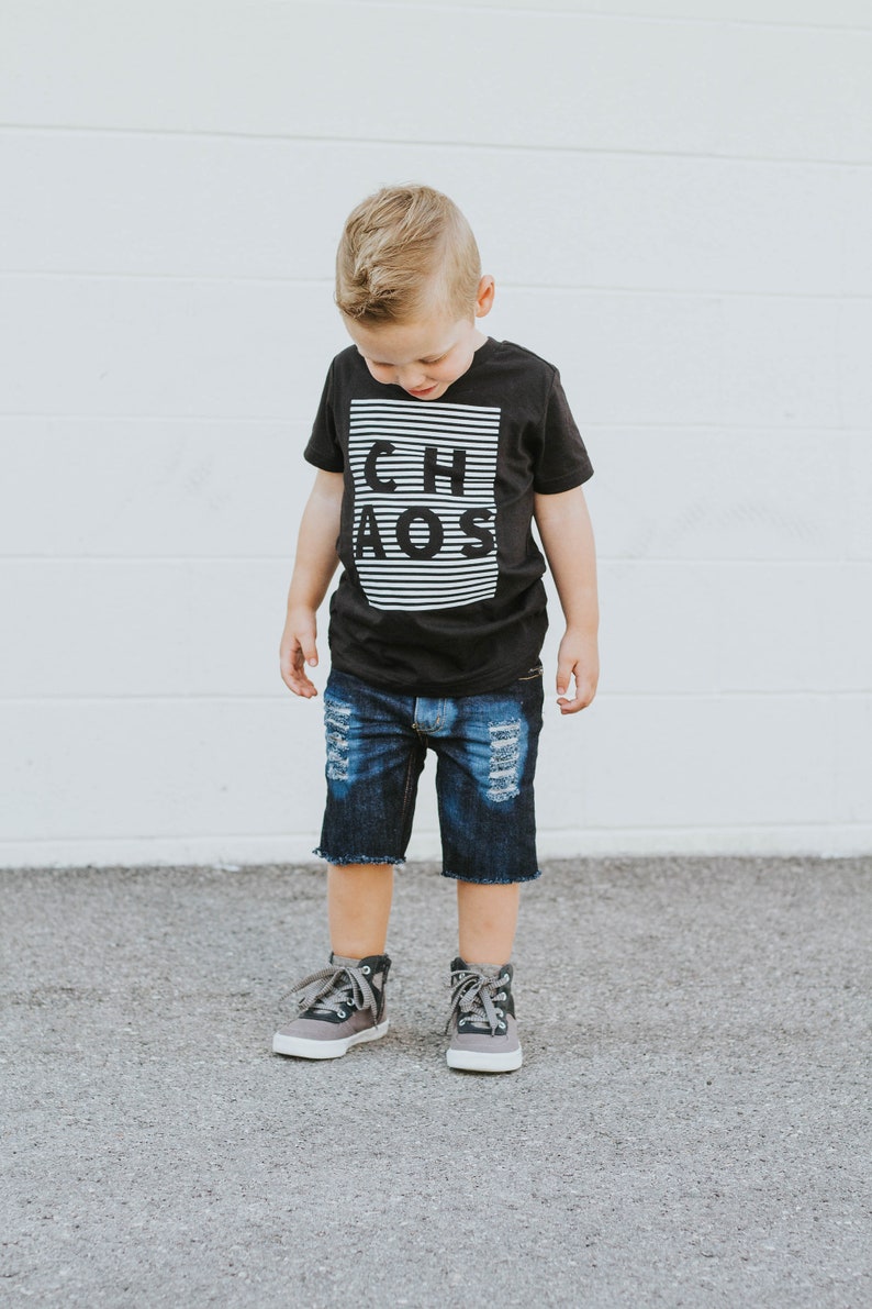 May include: A young boy wearing a black t-shirt with the word "CHAOS" printed in white on it. He is also wearing blue denim shorts and gray high-top sneakers.