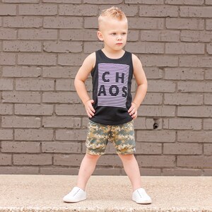 May include: A young boy wearing a black tank top with the word "CHAOS" printed on it in white letters. He is also wearing camouflage shorts and white sneakers.