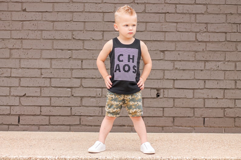 May include: A young boy wearing a black tank top with the word "CHAOS" printed on it in white letters. He is also wearing camouflage shorts and white sneakers.
