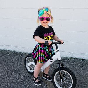May include: A young child wearing a black t-shirt with the text "Mama's Bro" in pink and yellow, a colourful tie-dye headband, and a black and white checked skirt, rides a white balance bike with black tyres.
