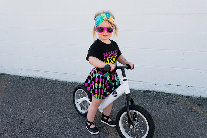 May include: A young child wearing a black t-shirt with the text "Mama's Bro" in pink and yellow, a colourful tie-dye headband, and a black and white checked skirt, rides a white balance bike with black tyres.