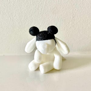 May include: A white plush toy in a seated position, featuring a black cap with two round ears. The toy has long, floppy ears and a small tail. The toy is sitting on a white surface.