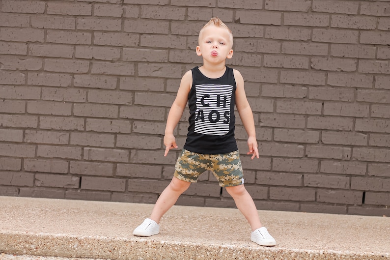 May include: A young boy with a spiky hairstyle wears a black tank top with white stripes and the word "CHAOS" printed on it. He is wearing camouflage shorts and white sneakers. He is standing in front of a brick wall and sticking his tongue out.