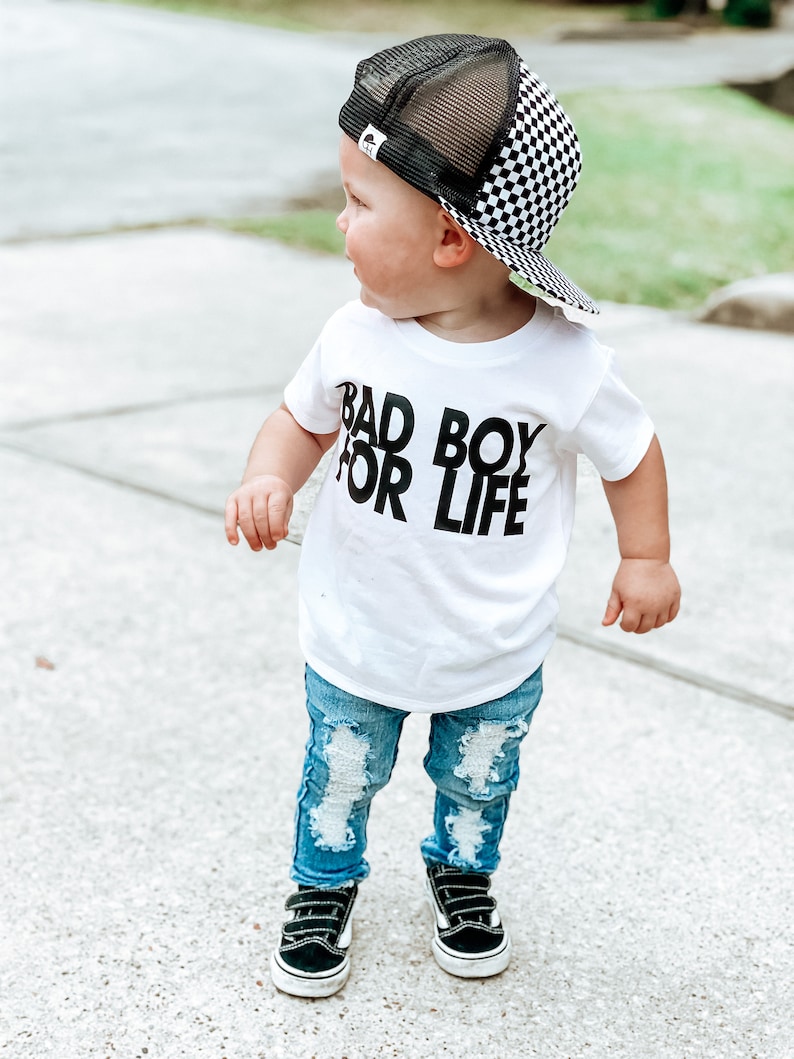 May include: A toddler wearing a white t-shirt with the text "Bad Boy For Life" in black, blue ripped jeans, black trainers, and a black and white chequered trucker hat.