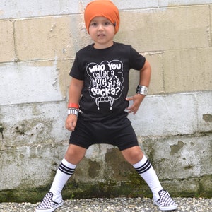 Pu&ograve; includere: Un bambino piccolo che indossa una maglietta nera con la scritta "Who you callin' a sucka sucka?" in bianco, pantaloncini neri, berretto arancione, calze bianche con strisce nere e scarpe da ginnastica a scacchi nere e bianche. Il bambino &egrave; in piedi davanti a un muro di mattoni bianchi.