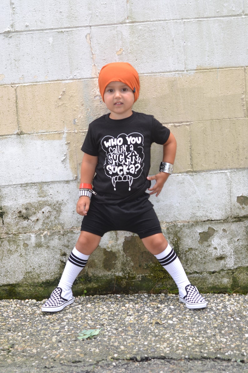 Pu&ograve; includere: Un bambino piccolo che indossa una maglietta nera con la scritta "Who you callin' a sucka sucka?" in bianco, pantaloncini neri, berretto arancione, calze bianche con strisce nere e scarpe da ginnastica a scacchi nere e bianche. Il bambino &egrave; in piedi davanti a un muro di mattoni bianchi.