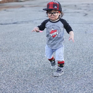 K&ouml;nnte beinhalten: Ein junges Kind tr&auml;gt ein grau-schwarzes Baseball-T-Shirt mit einem roten Herz-Motiv und dem Text "Ich brauche ein M&auml;dchen wie dich - Ich brauche ein M&auml;dchen wie dich - Mama". Es tr&auml;gt auch eine rot-schwarze karierte Baseballkappe, blaue Jeans-Shorts und schwarz-wei&szlig;e karierte Socken mit schwarz-wei&szlig;en karierten Turnschuhen.