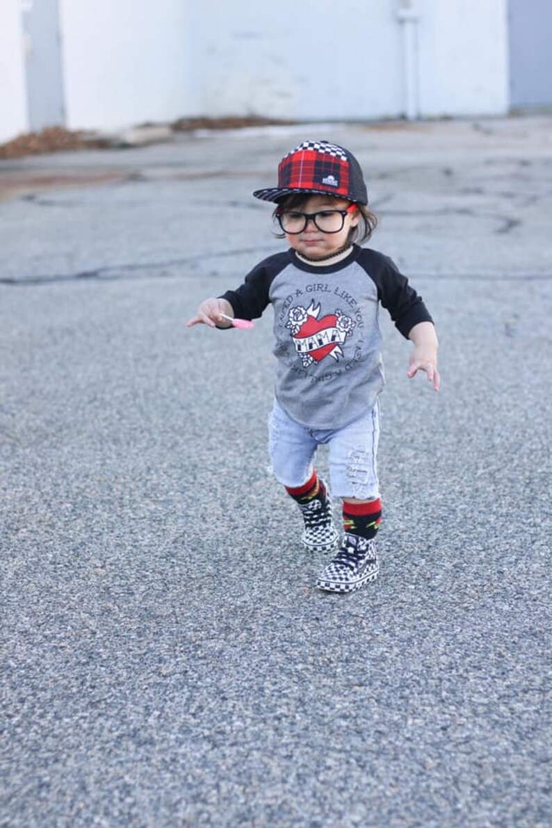 K&ouml;nnte beinhalten: Ein junges Kind tr&auml;gt ein grau-schwarzes Baseball-T-Shirt mit einem roten Herz-Motiv und dem Text "Ich brauche ein M&auml;dchen wie dich - Ich brauche ein M&auml;dchen wie dich - Mama". Es tr&auml;gt auch eine rot-schwarze karierte Baseballkappe, blaue Jeans-Shorts und schwarz-wei&szlig;e karierte Socken mit schwarz-wei&szlig;en karierten Turnschuhen.