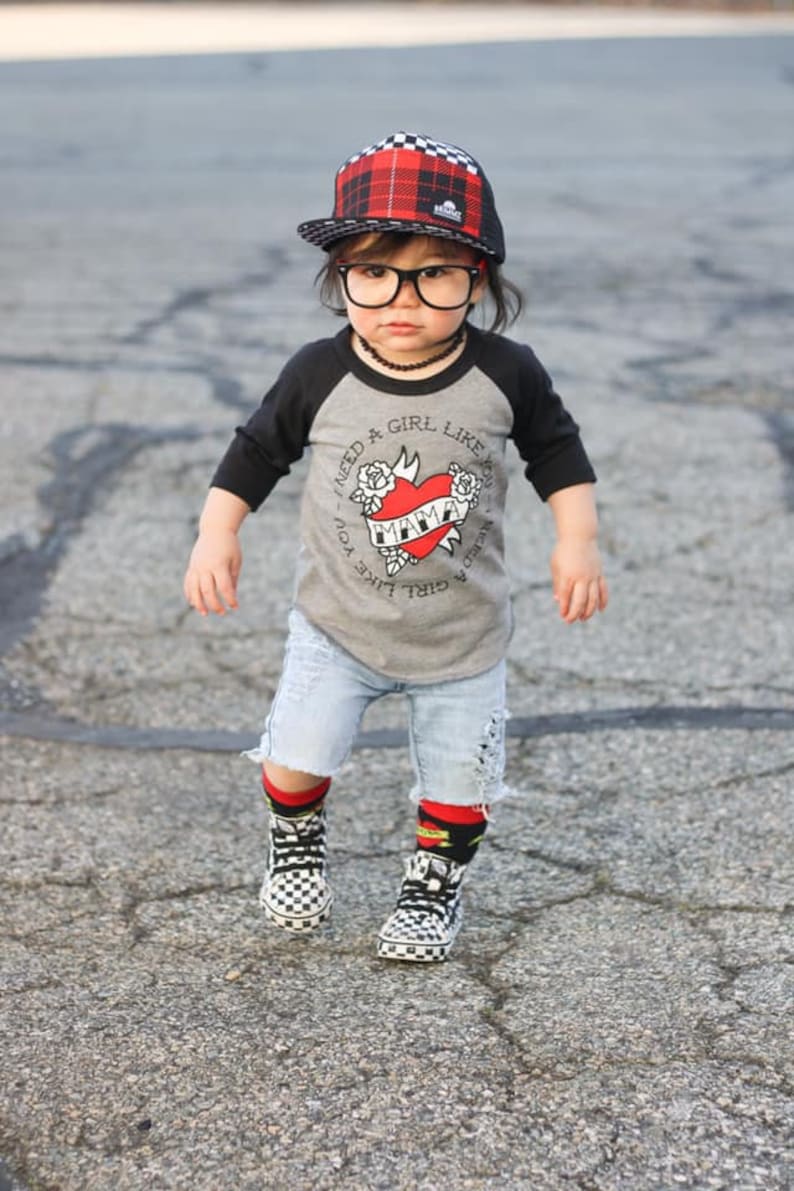 K&ouml;nnte beinhalten: Ein junges Kind tr&auml;gt ein grau-schwarzes Baseball-T-Shirt mit einem roten Herz-Motiv, auf dem "Ich brauche ein M&auml;dchen wie dich" und "Ich brauche ein M&auml;dchen wie dich" steht. Das Kind tr&auml;gt au&szlig;erdem eine schwarze und rot karierte Baseballkappe, eine schwarze Brille, eine blaue Jeans-Shorts und schwarz-wei&szlig; karierte Socken mit schwarz-wei&szlig; karierte Schuhen.