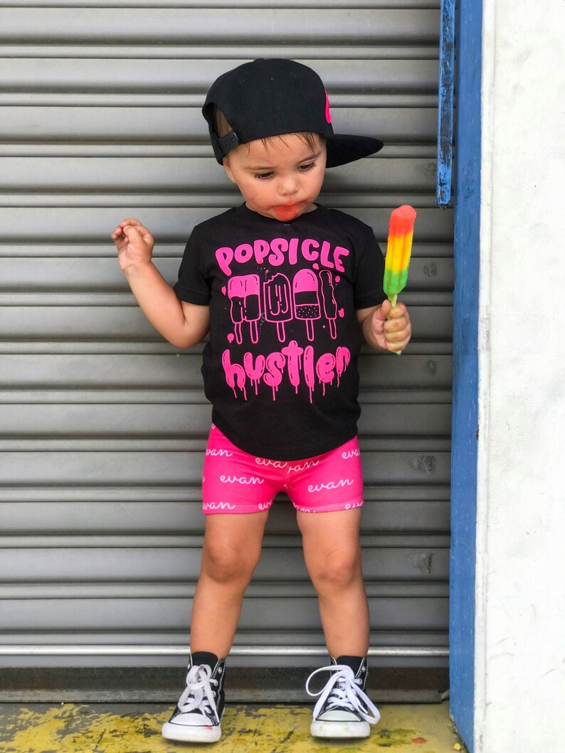 May include: A young child wearing a black t-shirt with the words "Popsicle Hustler" and a pink popsicle graphic. The child is also wearing a black baseball cap, pink shorts with a white pattern, and black and white sneakers. The child is holding a rainbow popsicle.