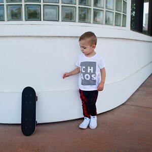 May include: A young boy wearing a white t-shirt with the text "Little Boss" printed on it, red and black plaid pants, and white sneakers. He is standing next to a black skateboard.