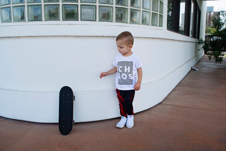 May include: A young boy wearing a white t-shirt with the text "Little Boss" printed on it, red and black plaid pants, and white sneakers. He is standing next to a black skateboard.