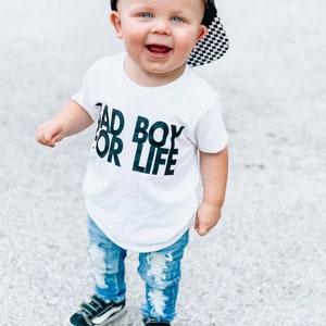 May include: A toddler wearing a white t-shirt with the text "Bad Boy For Life" in black, blue ripped jeans, black trainers, and a black and white chequered baseball cap.