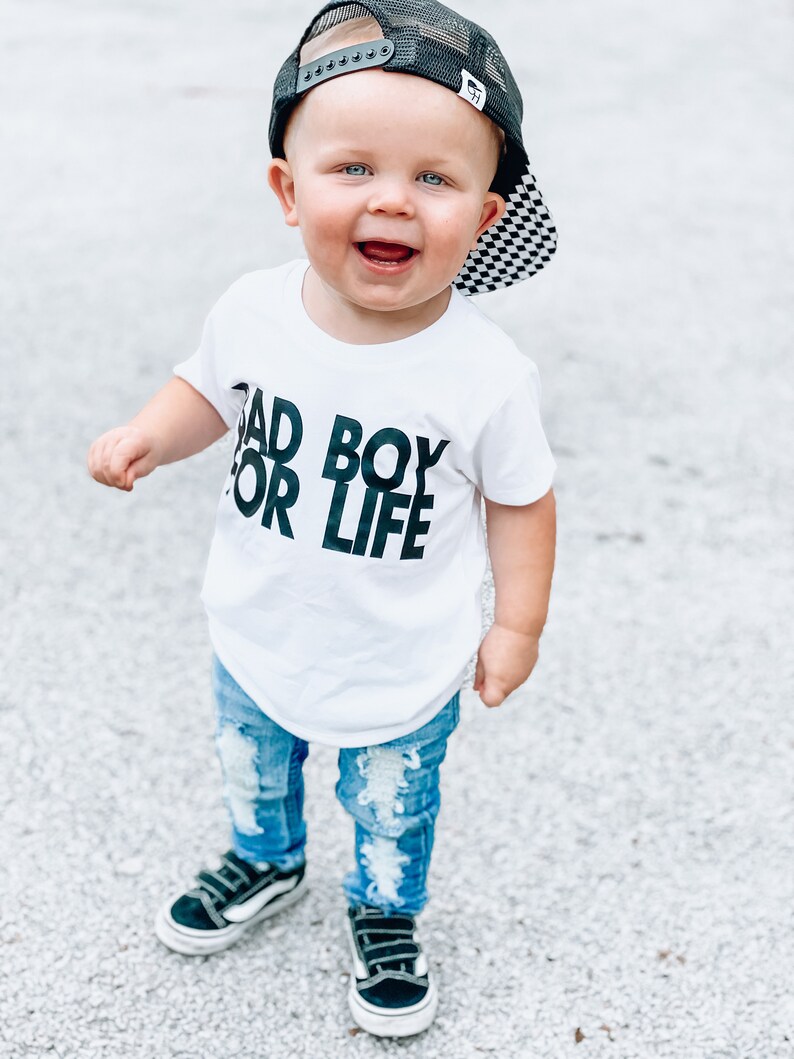 May include: A toddler wearing a white t-shirt with the text "Bad Boy For Life" in black, blue ripped jeans, black trainers, and a black and white chequered baseball cap.