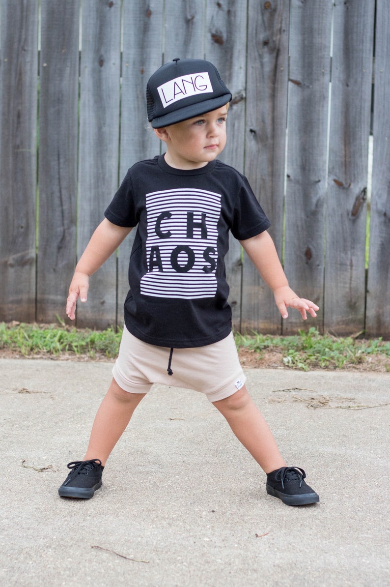 May include: A young person wearing a black t-shirt with the word "CHAOS" printed on it, beige shorts, black sneakers, and a black mesh baseball cap with the word "LANG" printed on it.