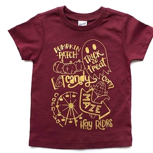 Może przedstawiać: Bordowy t-shirt z złotym nadrukiem przedstawiającym pole z dyniami, kukurydzę, ducha i tekst "Pumpkin Patch, Trick or Treat, Candy Corn, Carnival, Maze, Hay Rides".
