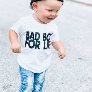 May include: A toddler wearing a white t-shirt with the text "Bad Boy For Life" in black lettering, blue ripped jeans, black trainers, and a black mesh trucker hat.