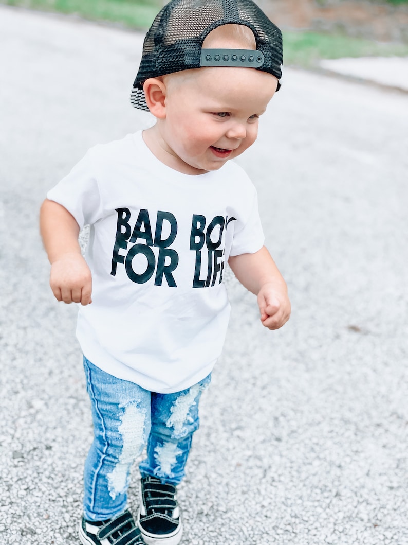 May include: A toddler wearing a white t-shirt with the text "Bad Boy For Life" in black lettering, blue ripped jeans, black trainers, and a black mesh trucker hat.