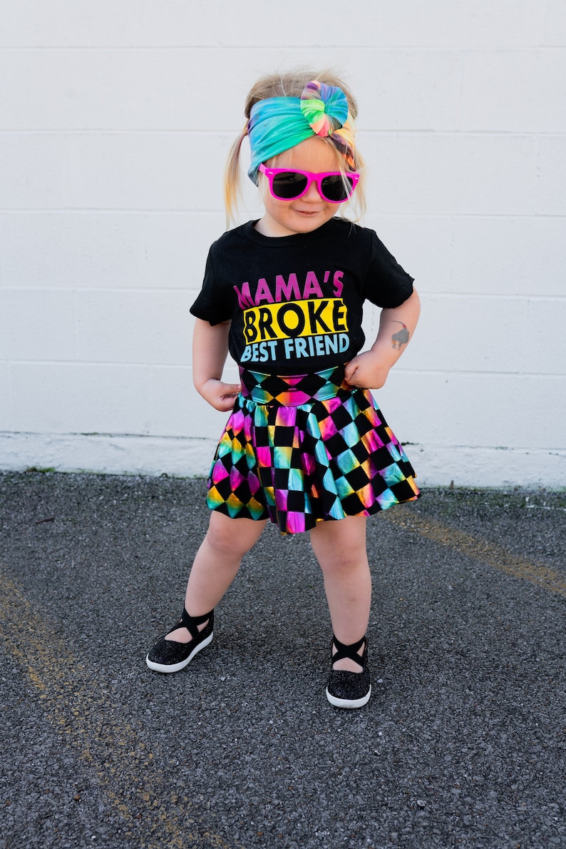 May include: A young person wearing a black t-shirt with the text "Mama's Broke Best Friend" in pink, yellow, and blue. They are also wearing a rainbow tie-dye headband, pink sunglasses, a black and iridescent checkered skirt, and black sparkly shoes.