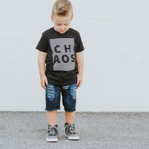 May include: A young boy wearing a black t-shirt with white stripes and the word "CHAOS" printed on it. He is also wearing blue denim shorts with distressed details and gray high-top sneakers.